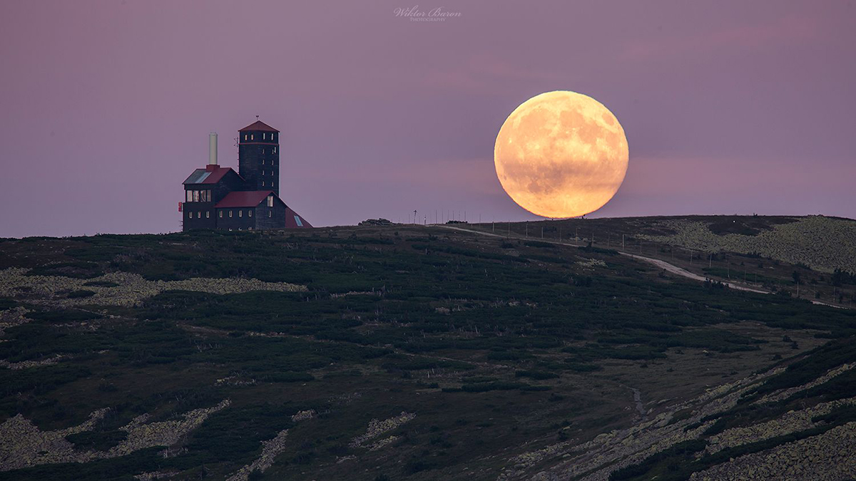 Large orange moon rising behind a mountaintop building at dusk.