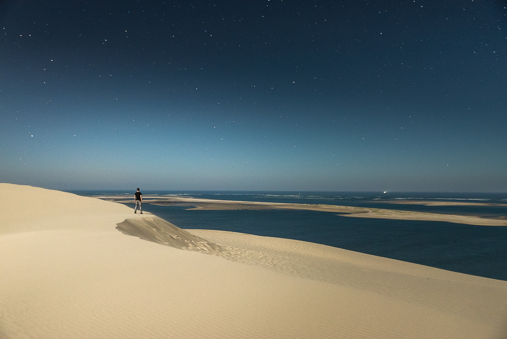Lone person stands on a pale sand dune beside the sea at twilight.