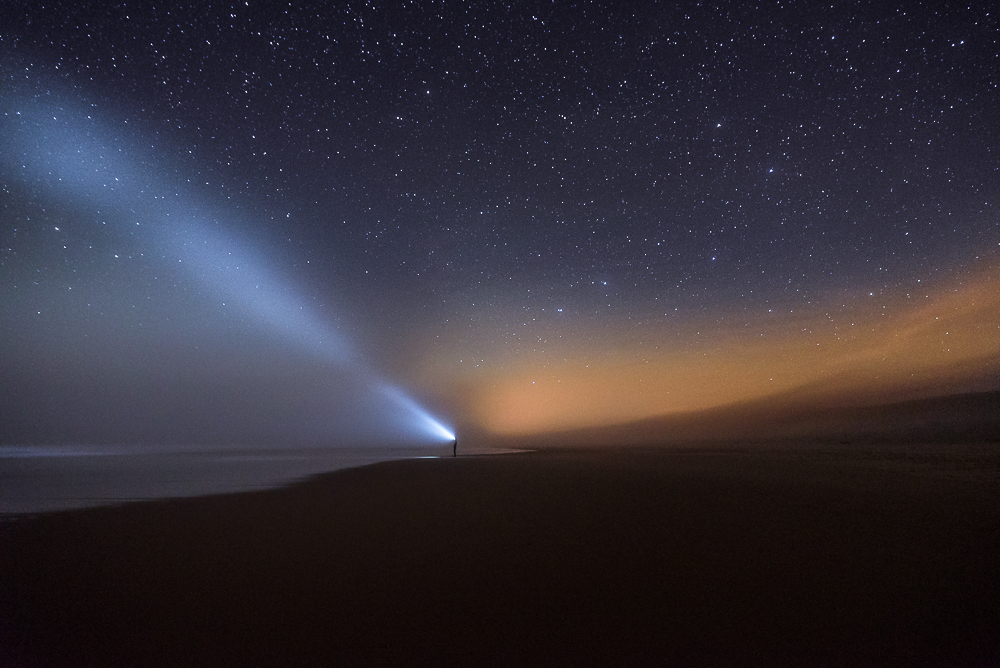 A beam of light cuts across a dark beach below a starry sky.