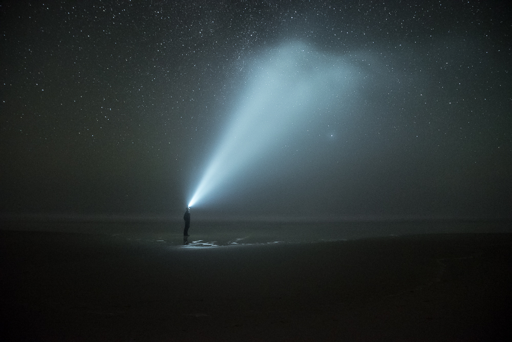 A beam of light from a lone figure sweeps across a dark beach under a starry sky.