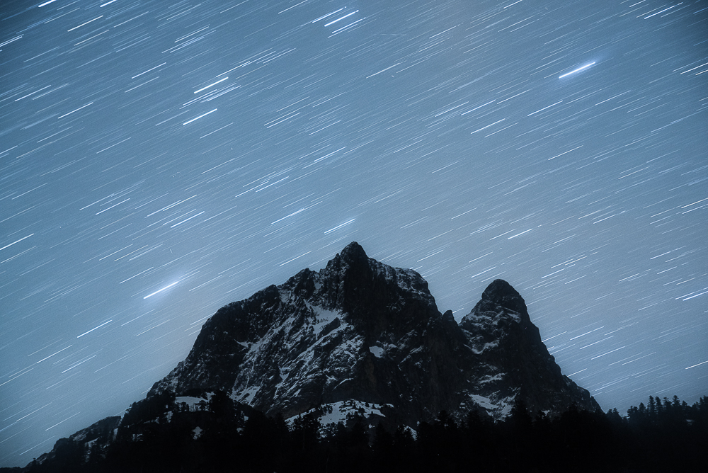 Star trails circle above silhouetted mountain peaks.