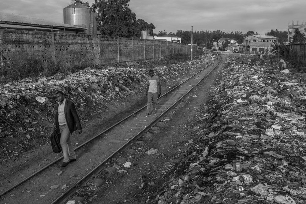 People walking beside railway tracks lined with rubbish and small buildings.