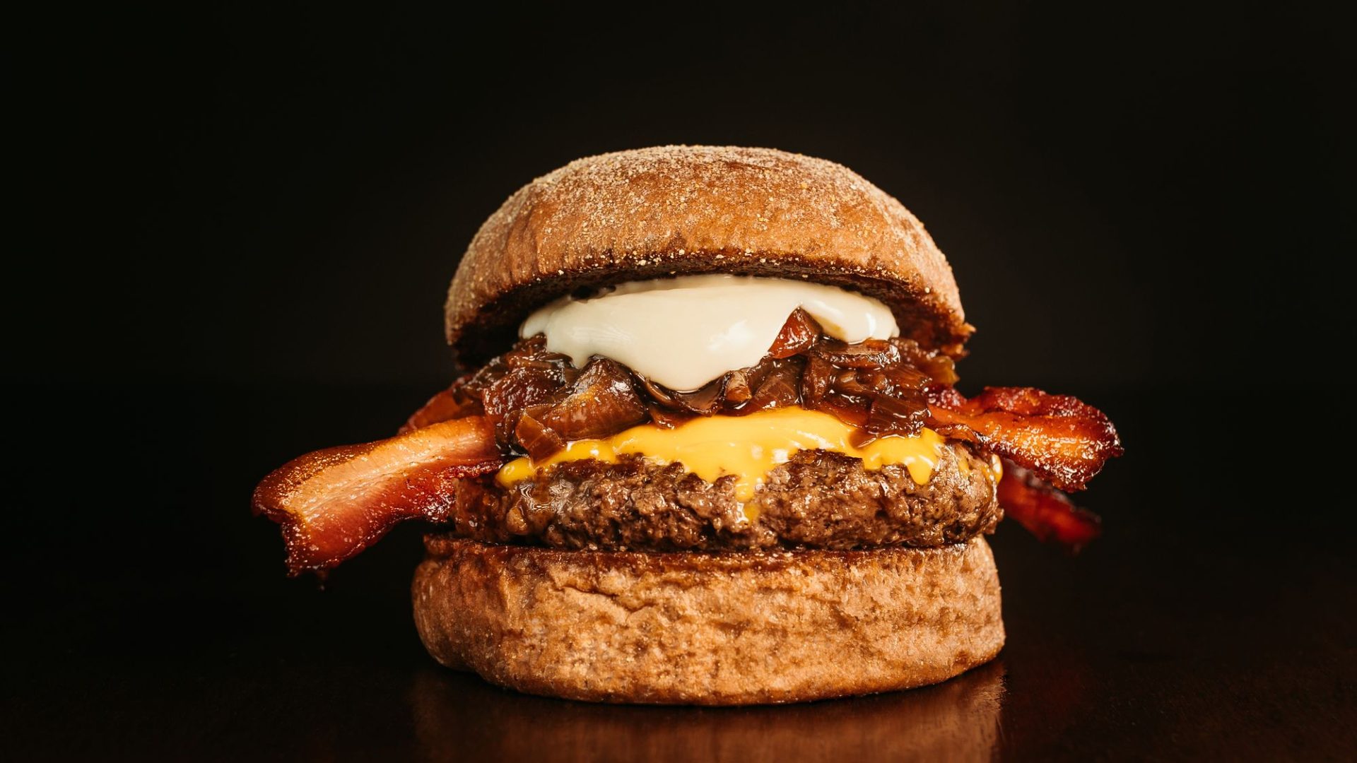 Close-up food photograph of a bacon cheeseburger with melted cheese and caramelized onions against a dark background