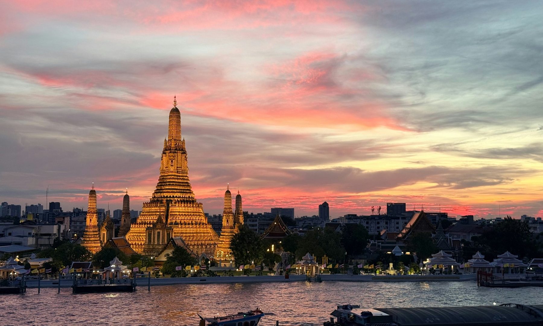 Wat Arun illuminated beside the Chao Phraya River under a pink sunset sky in Bangkok