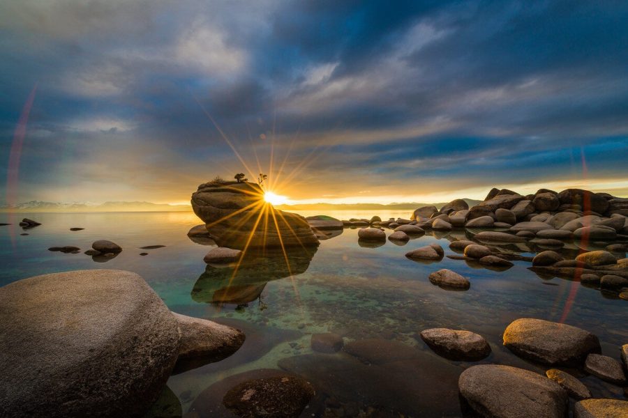 A sunburst shines beside Bonsai Rock at Lake Tahoe, reflected in calm water with foreground stones.
