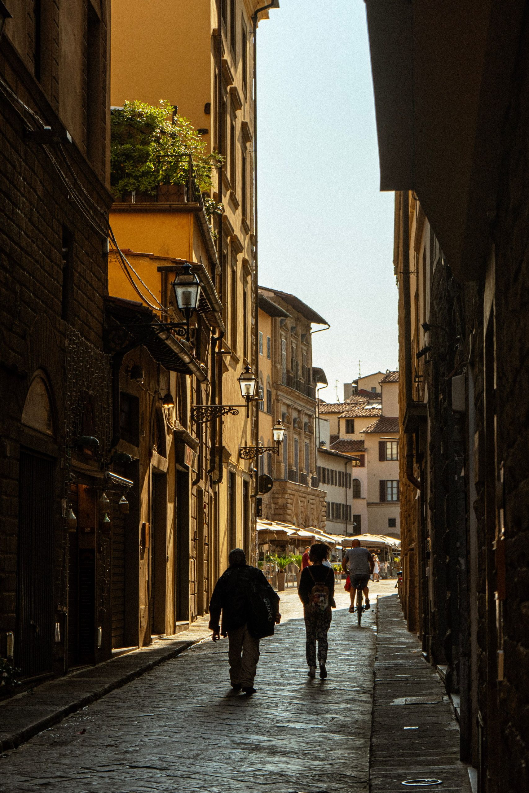 Two people walking away from the camera down a narrow, sunlit Florence street between tall stone buildings.
