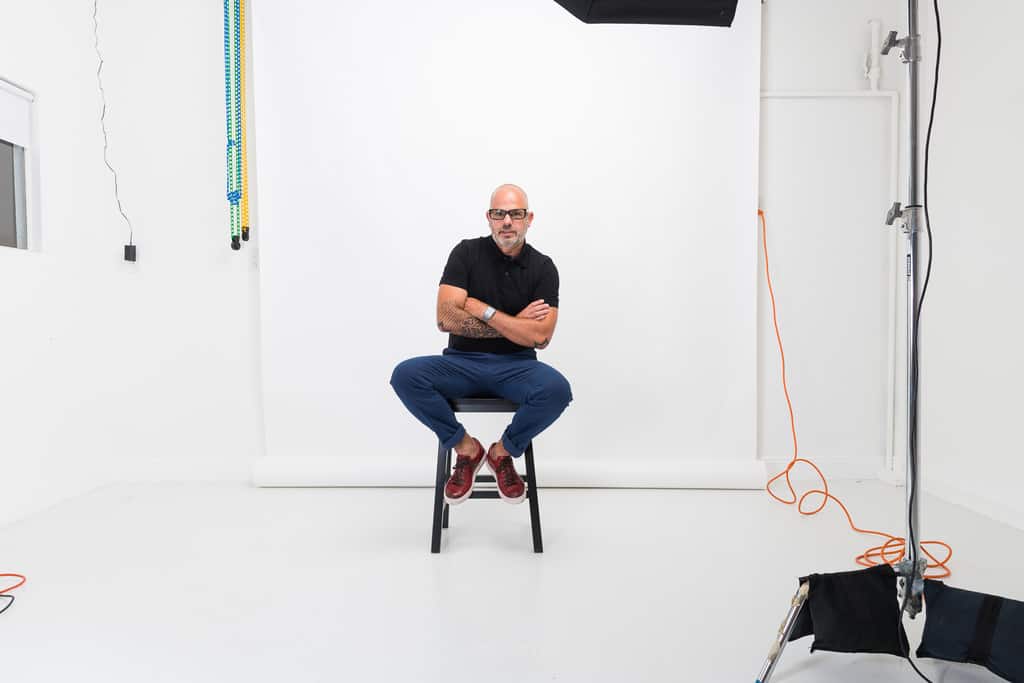 Seated man in a white studio with backdrop and lighting equipment visible