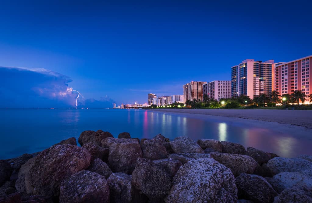 Lightning striking the ocean off Miami Beach at blue hour, with glowing shoreline hotels behind a rocky foreground