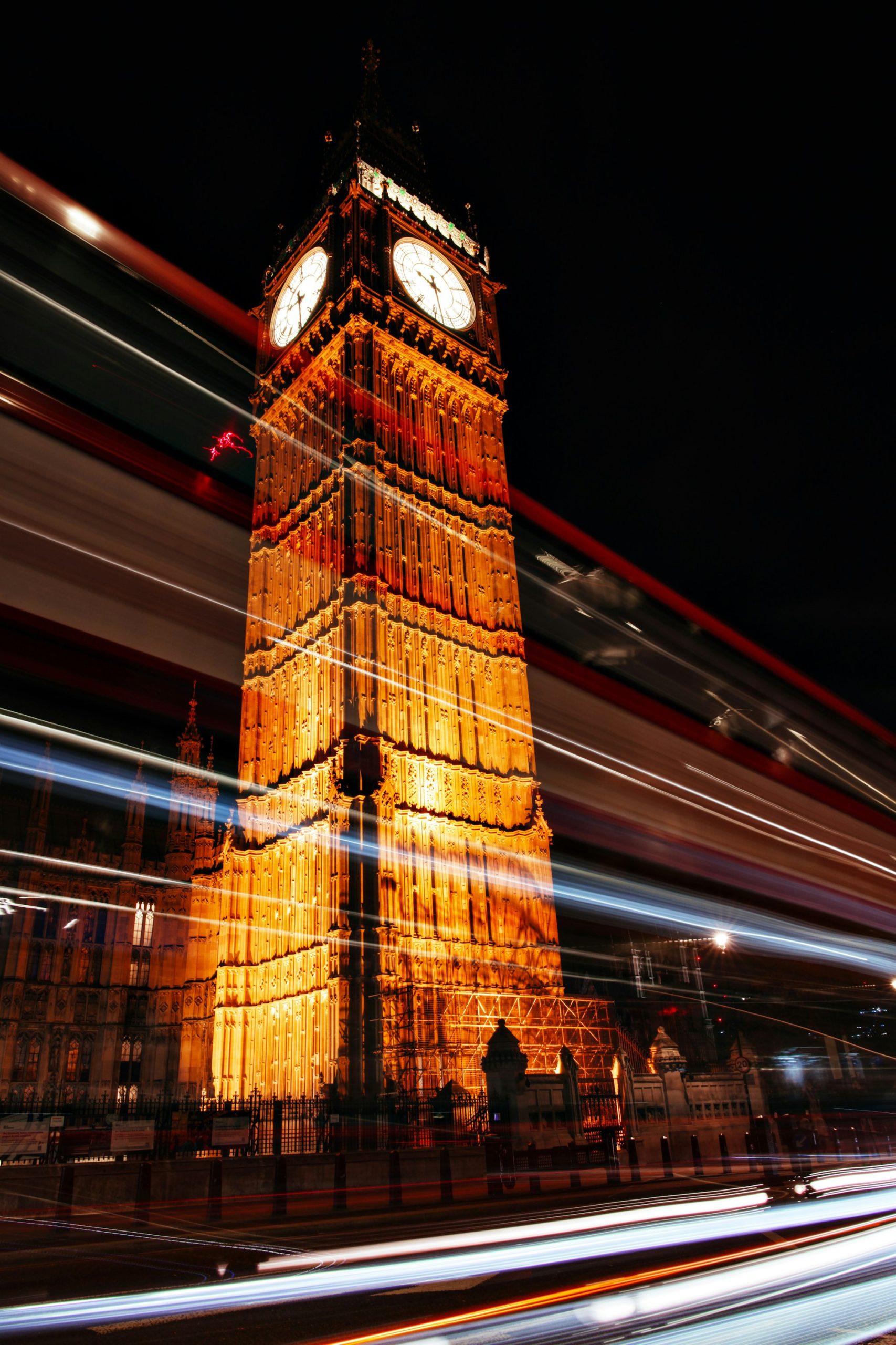 Big Ben (Elizabeth Tower) glowing gold at night beside streaking red and blue light trails from passing London buses