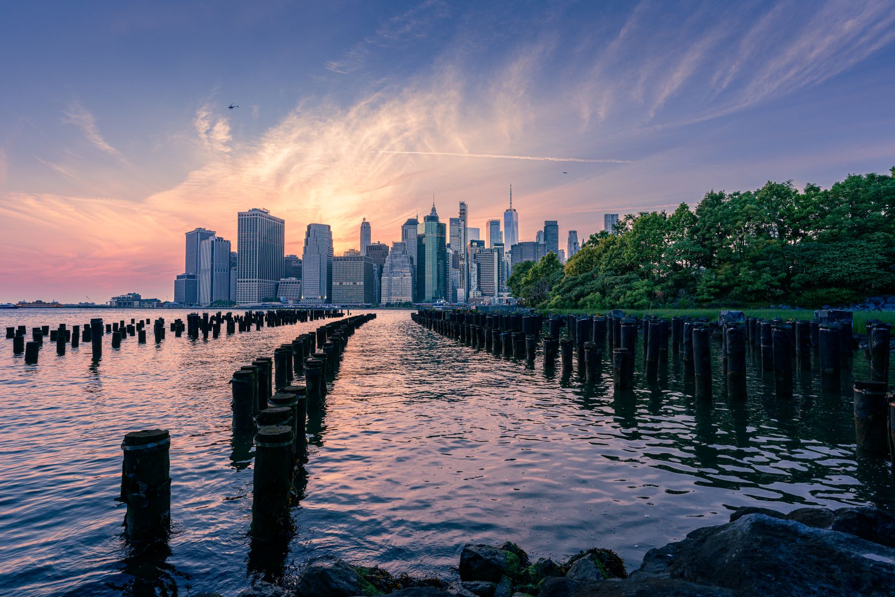 The Manhattan skyline at sunset seen across the East River from Brooklyn Bridge Park, with old wooden pier posts in the water and a pink and purple sky