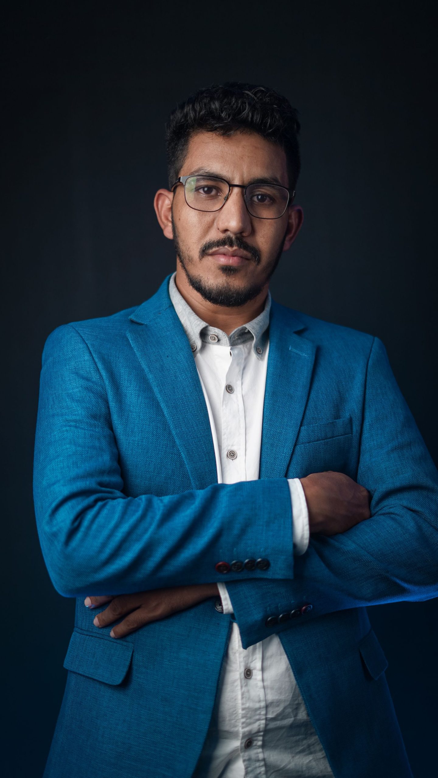 Man in a blue blazer and white shirt with his arms crossed against a dark studio background