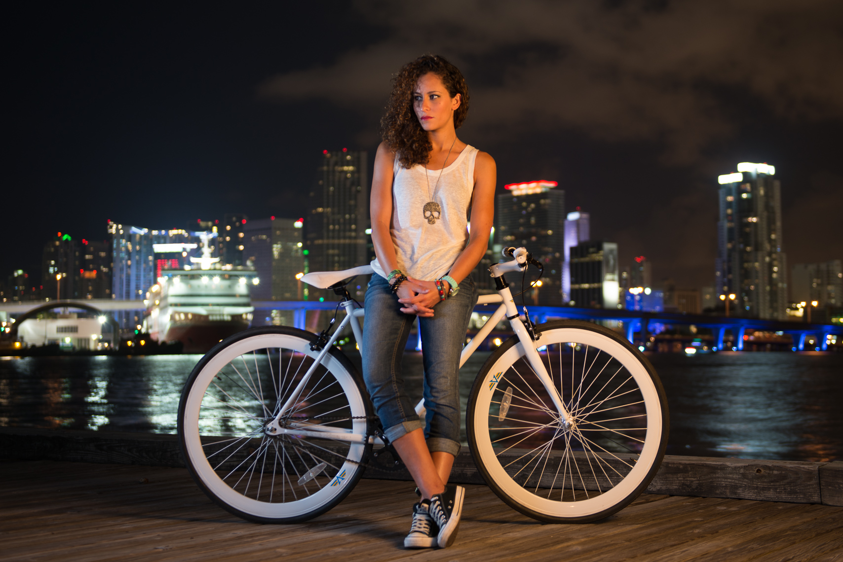 The before frame: night portrait of a woman with a white bicycle on the Miami waterfront, warm tones and city lights behind her