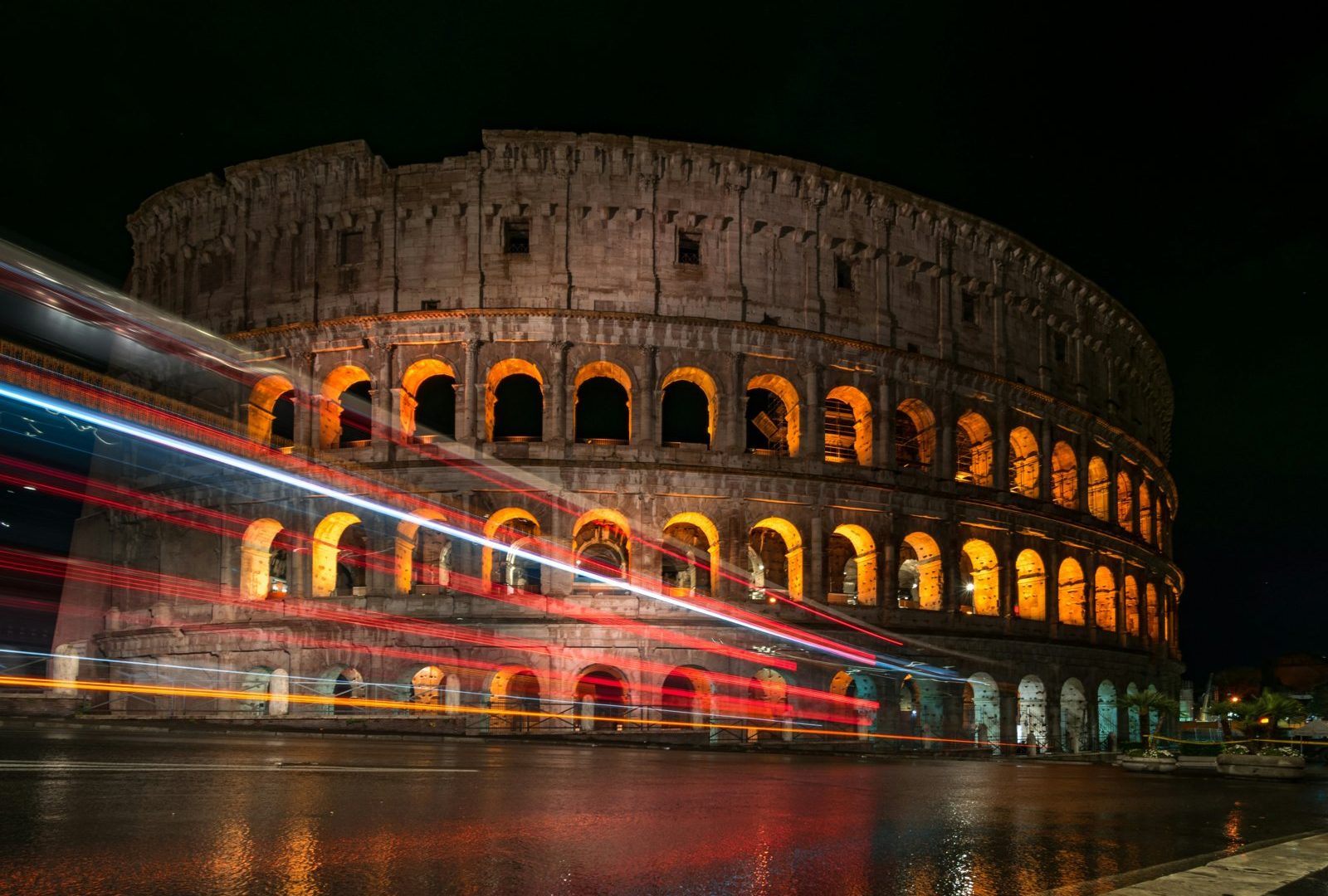 The Colosseum in Rome at night with orange-lit arches and light trails from passing traffic