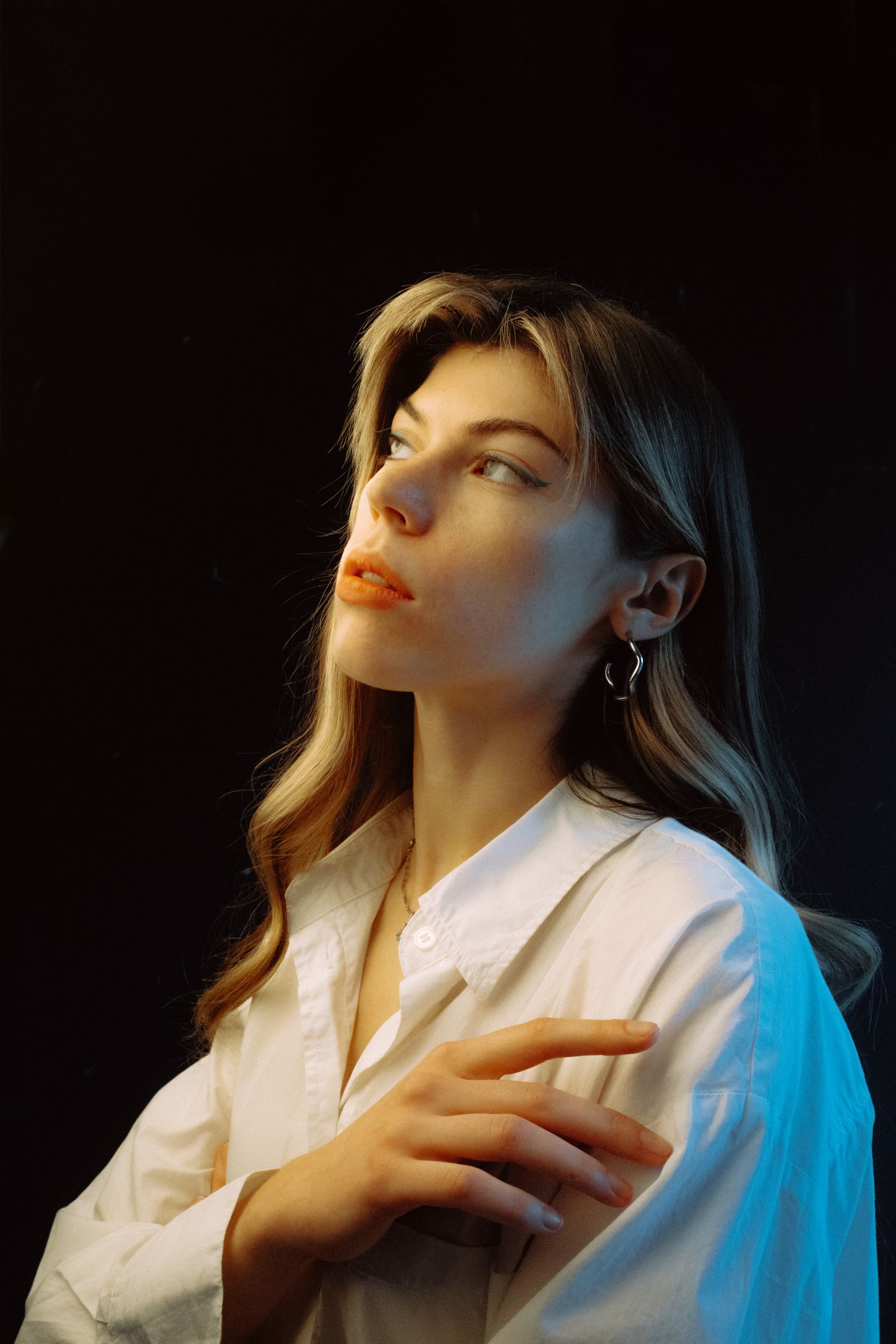 Woman in a white shirt looking away from the camera in a dark studio, lit from one side with warm and cool light on her face.