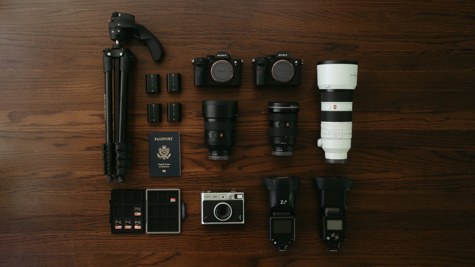 Flat lay of two mirrorless camera bodies, lenses, a tripod, batteries and memory cards on a wooden floor
