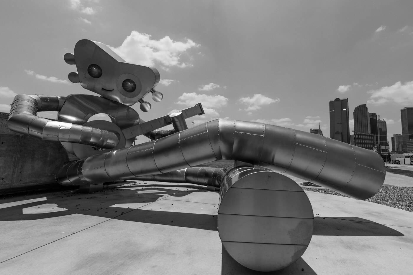 A black and white photo of a metal sculpture of a figure lying on its side, with city buildings behind it