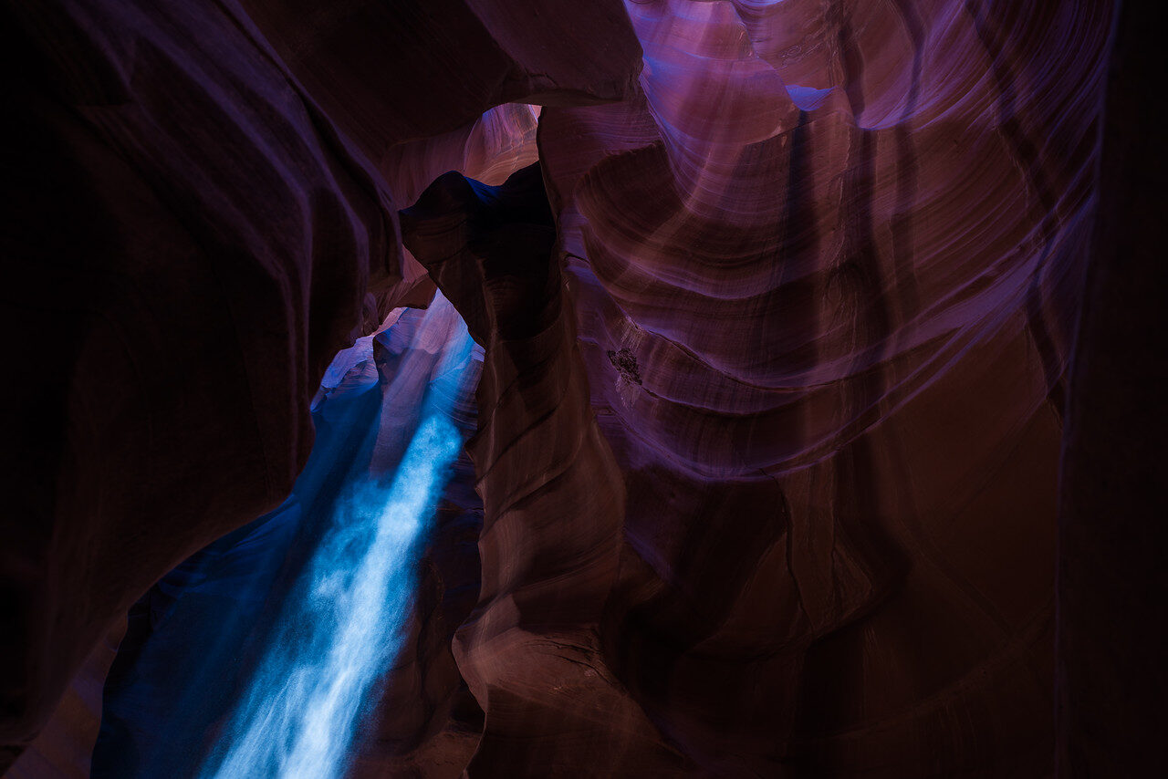 Blue shaft of light falling through a narrow slot canyon with purple and orange sandstone walls in Antelope Canyon, Arizona, photographed by Edin Chavez