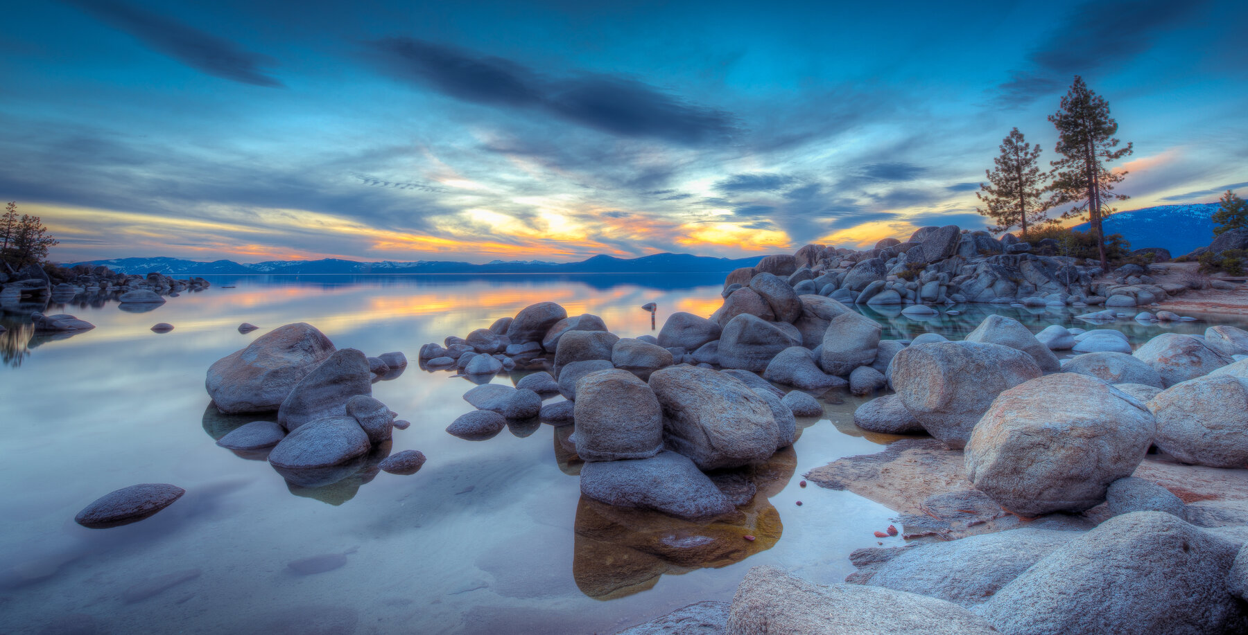 Rocks along a calm shoreline reflecting a blue sky with streaky clouds and distant mountains