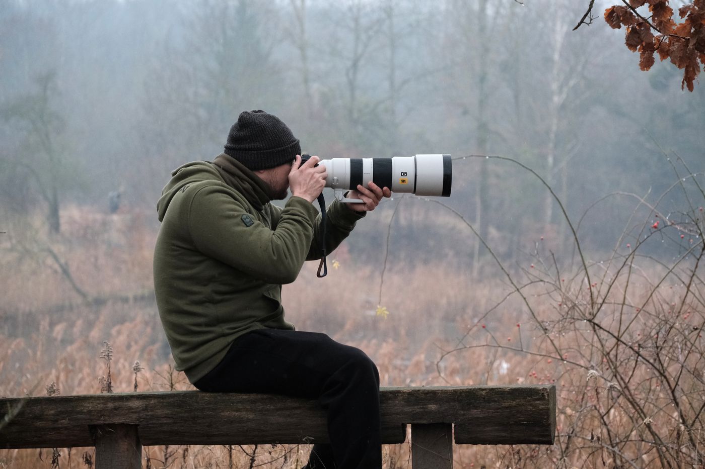 A photographer in a green jacket sitting on a bench holding a camera with a large white telephoto lens in misty woodland.