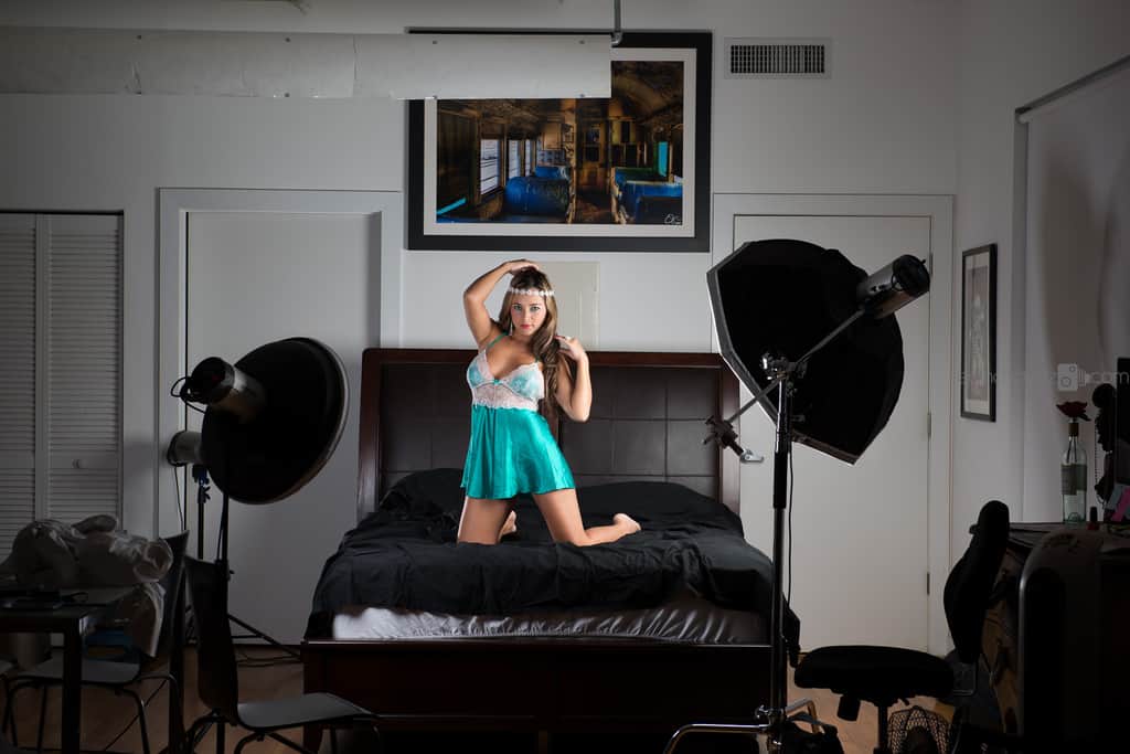 Wide view of a bedroom studio set with a black-bedded bed, a woman in a teal outfit kneeling on it, framed art on the wall and photography lighting equipment on stands around the room
