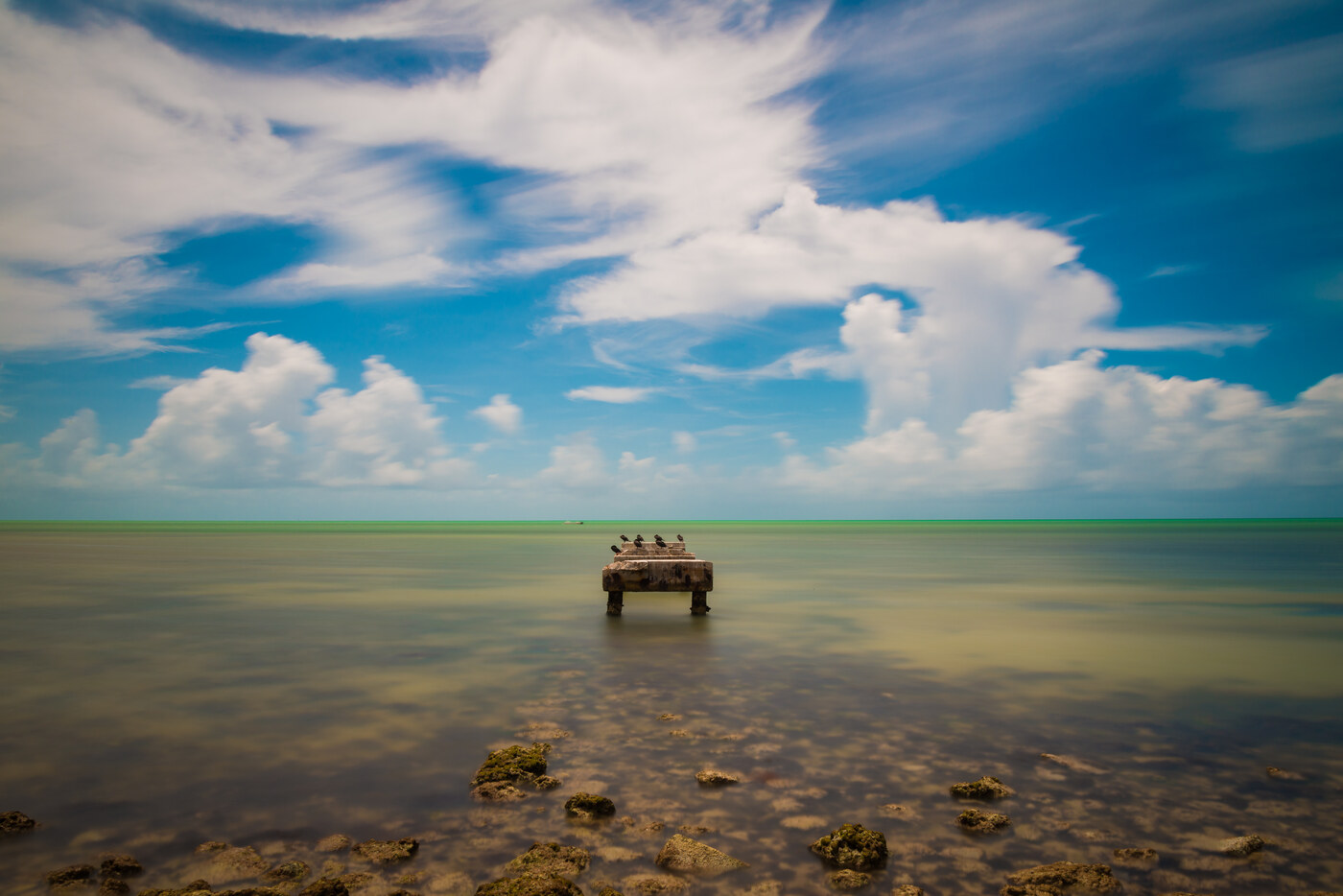 Bright white clouds above calm shallow water, with a few birds standing on a small wooden platform near the horizon