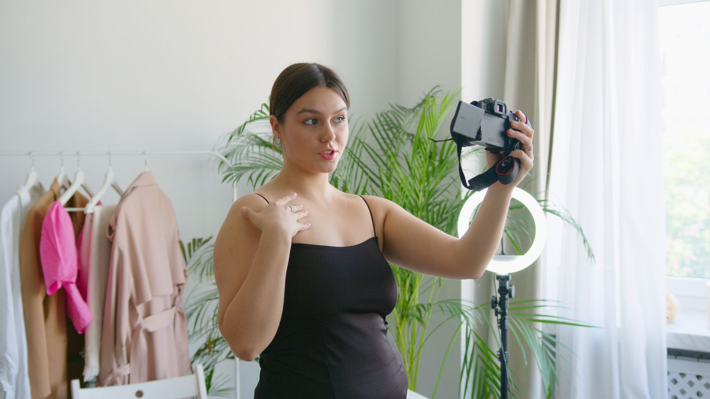 A woman holding a camera at arm's length in a bright room with a ring light on a stand behind her.
