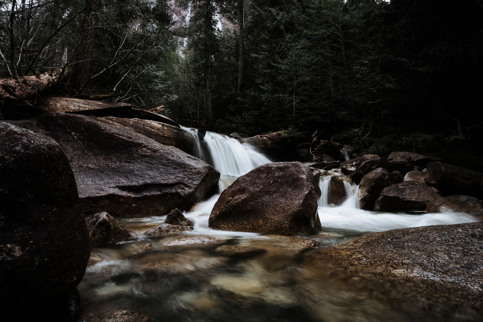 Long exposure of a forest stream flowing between large dark boulders with soft white water
