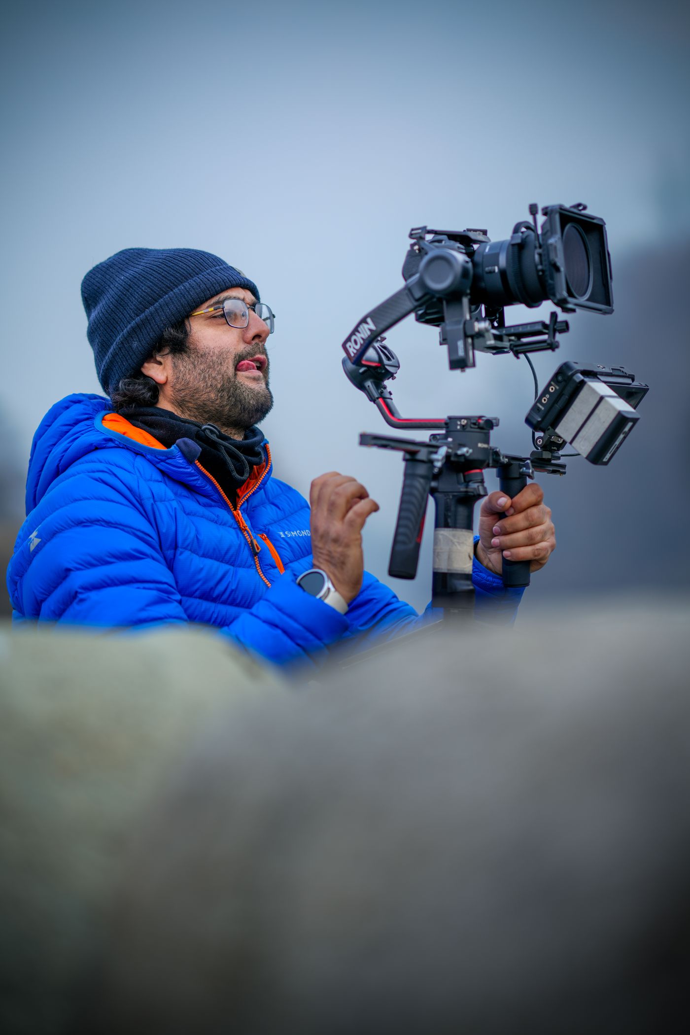 A man in a blue jacket and knit cap holding a camera on a handheld gimbal rig.