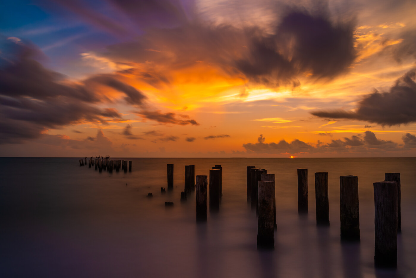 Rows of weathered wooden posts in smooth water under an orange and purple sunset sky