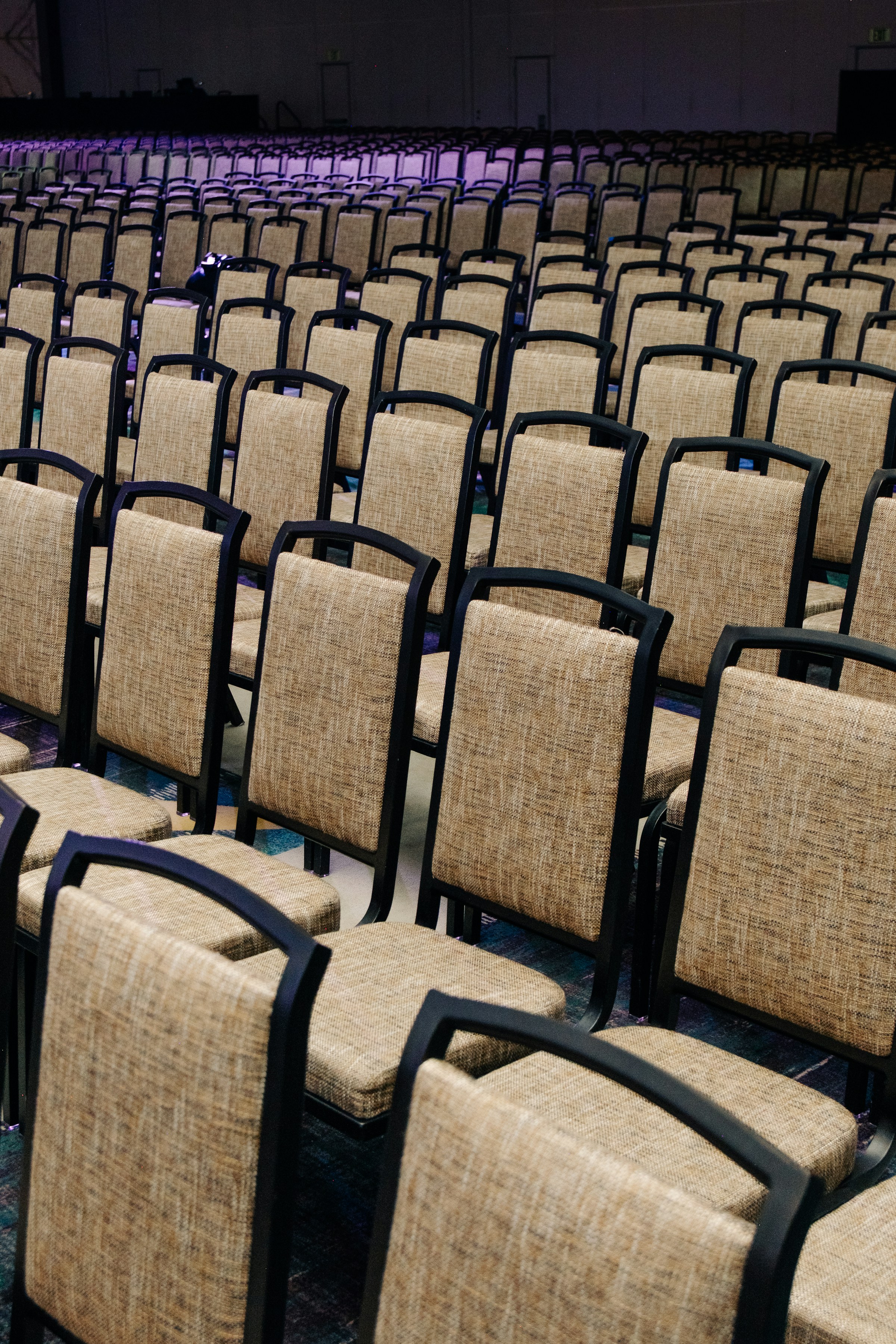 Rows of empty tan upholstered banquet chairs in a large hall with dark walls behind them