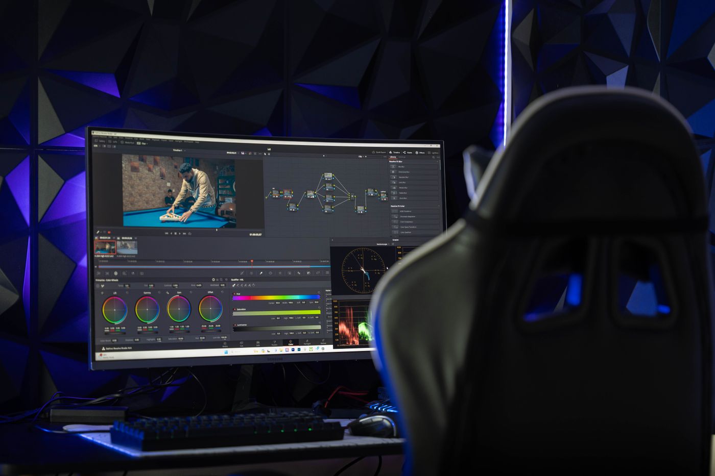 A computer monitor showing video editing and color grading software on a desk with a gaming chair beside it.