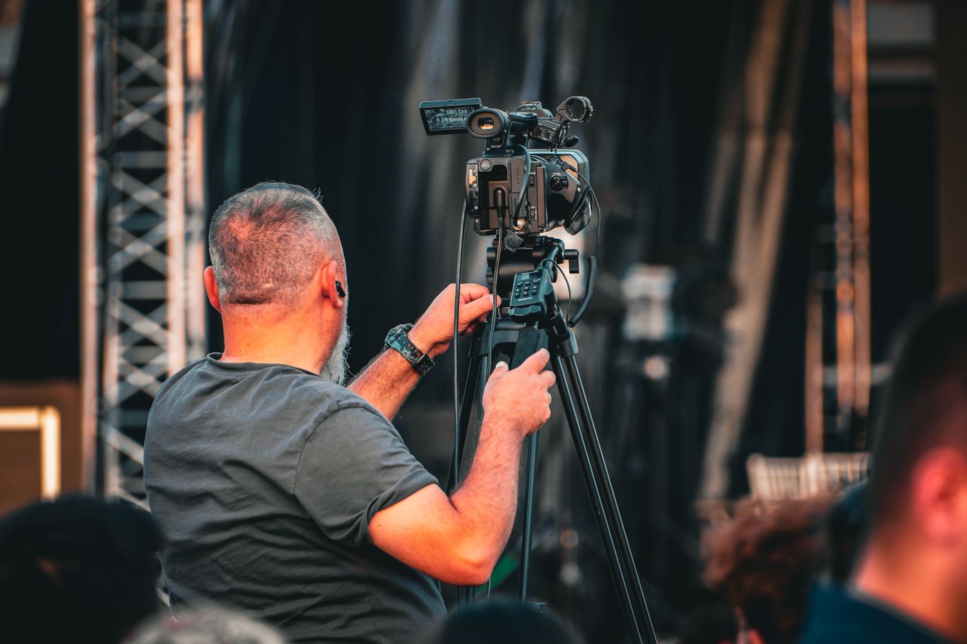 A camera operator in a gray shirt filming with a video camera on a tripod in front of a stage.