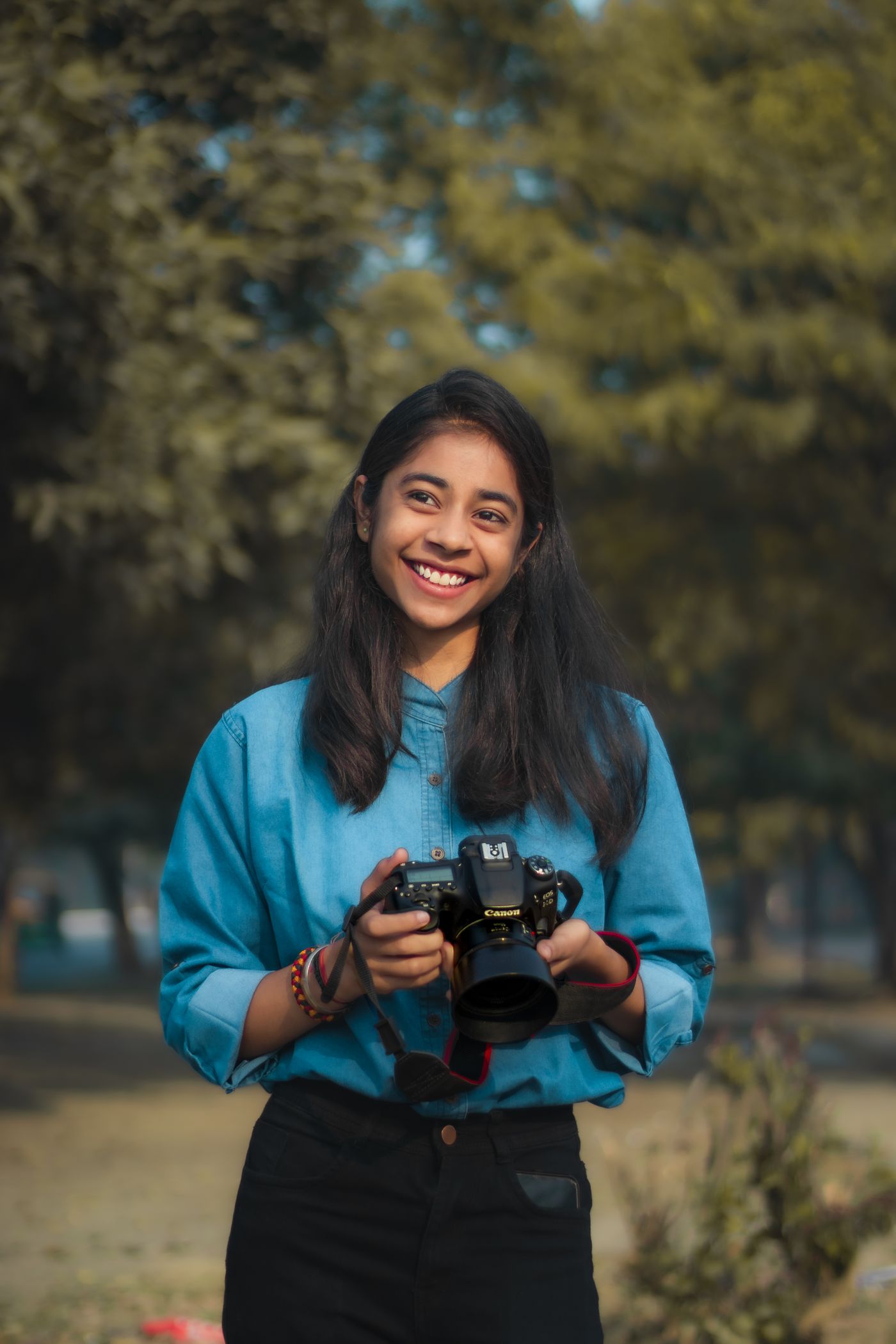 A smiling young woman in a blue shirt holding a camera with both hands in a park.