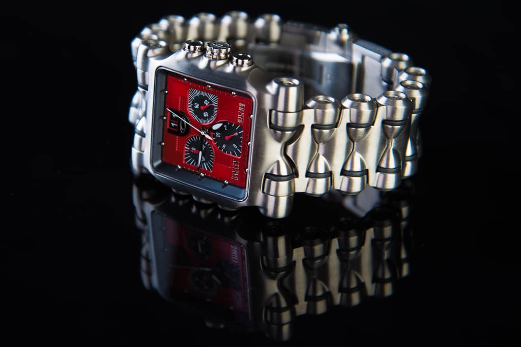 A wristwatch with a red dial on a glossy black reflective surface, lit like a studio product shot