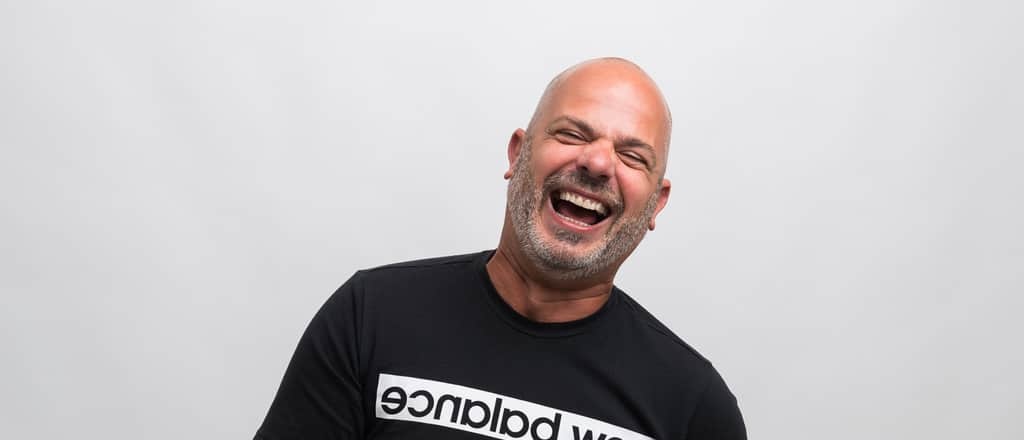 Laughing man in a black shirt against a light gray studio background.