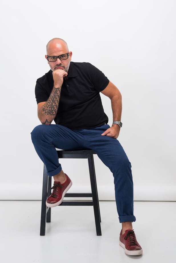 Jorge leaning forward on a stool with one hand near his chin in a full-length portrait
