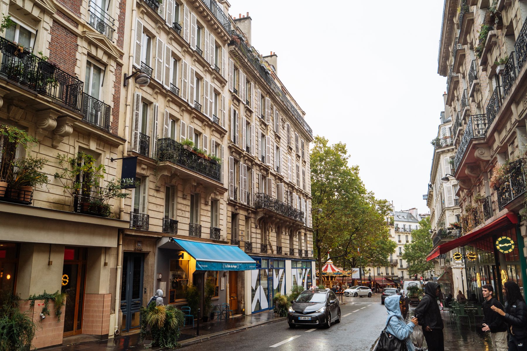 A rainy Paris street lined with cream Haussmann apartment buildings, wrought iron balconies and a parked car