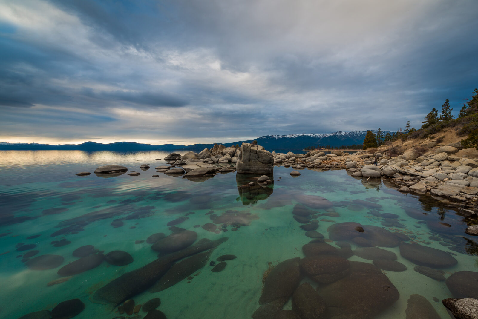 Clear green water and rounded granite boulders at Lake Tahoe under heavy clouds with a distant snowy ridge
