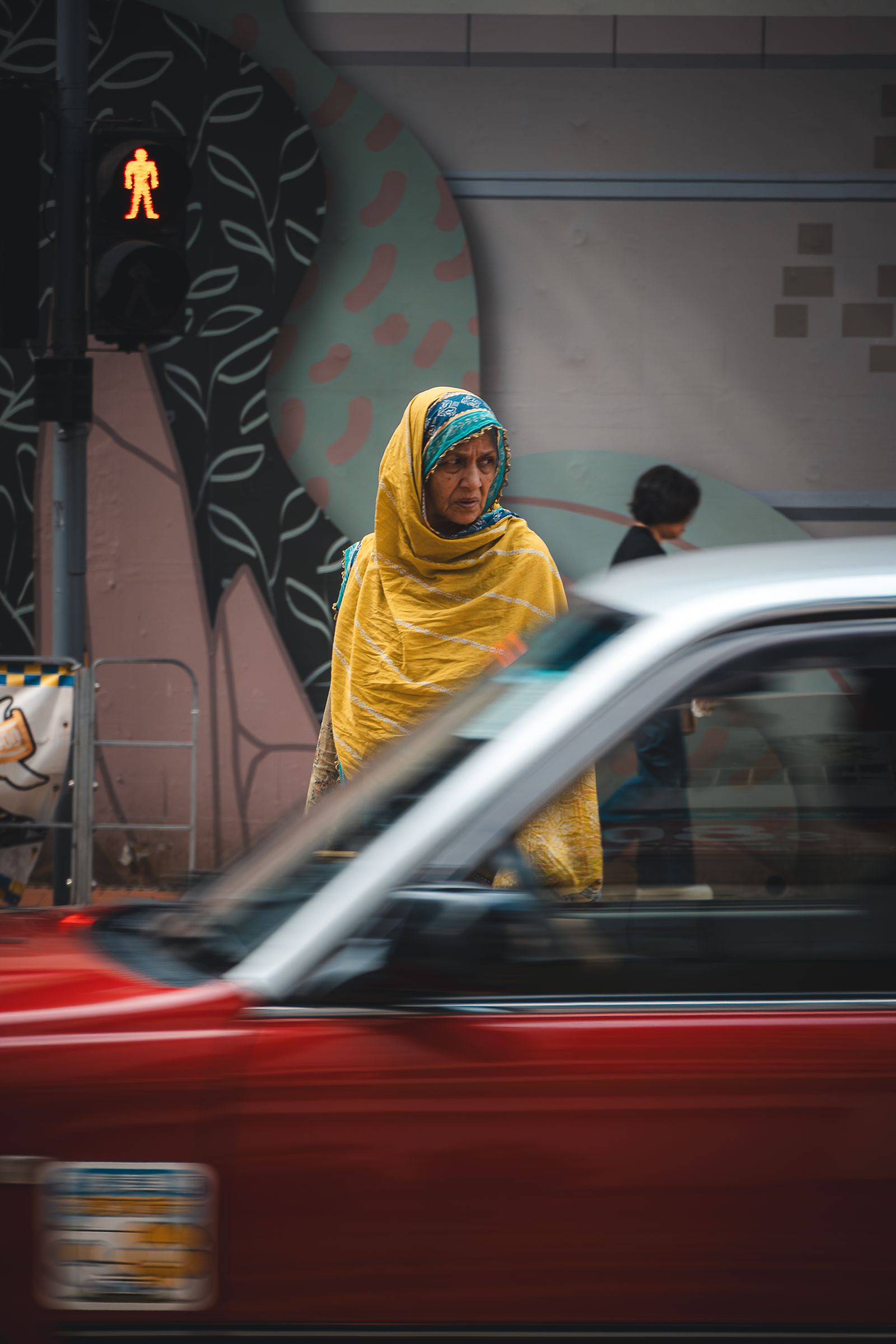 Woman in a yellow headscarf waits at a crossing as a red car blurs past in front of her