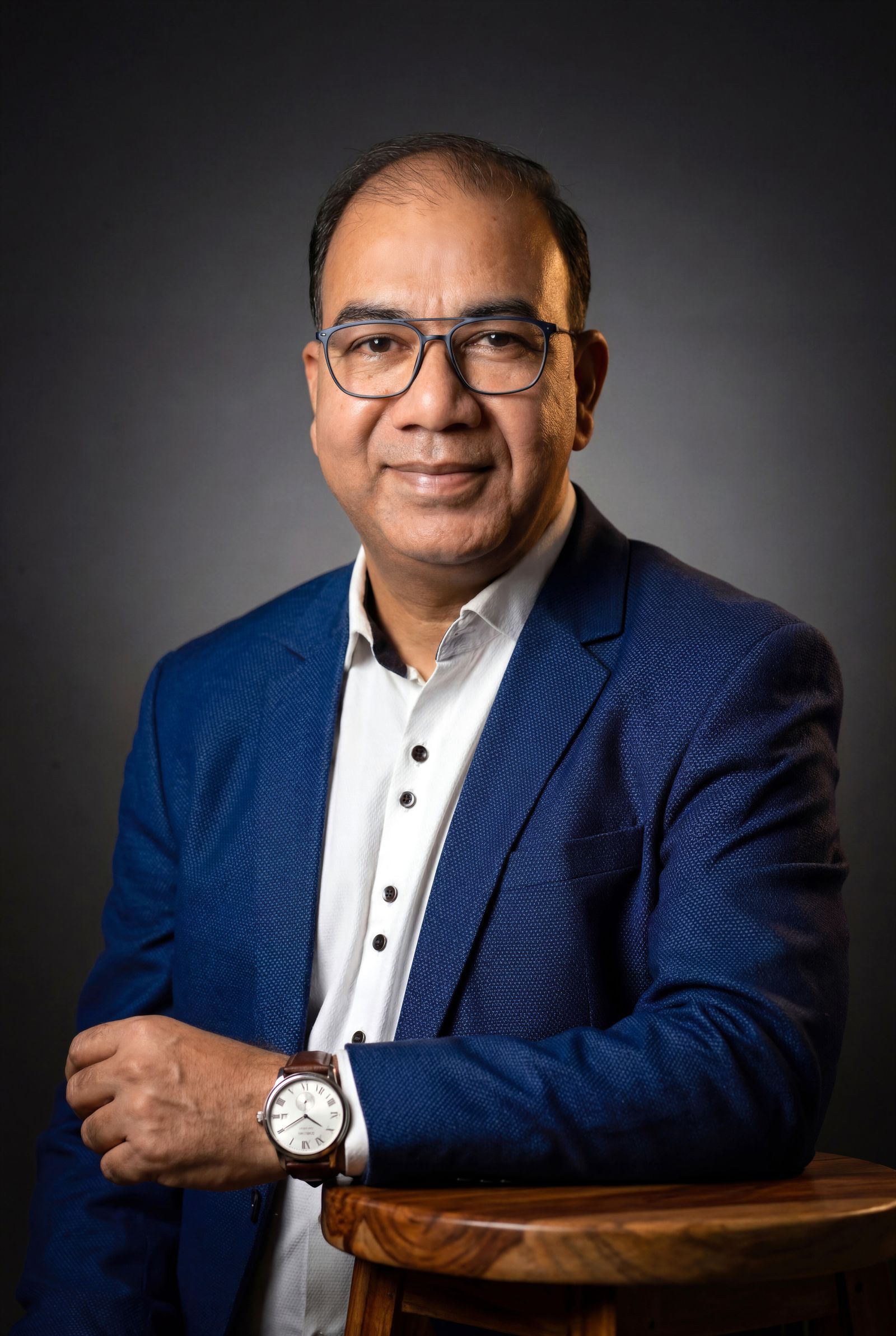 Smiling man with glasses in a blue blazer resting his arm on a wooden stool against a gray studio backdrop.