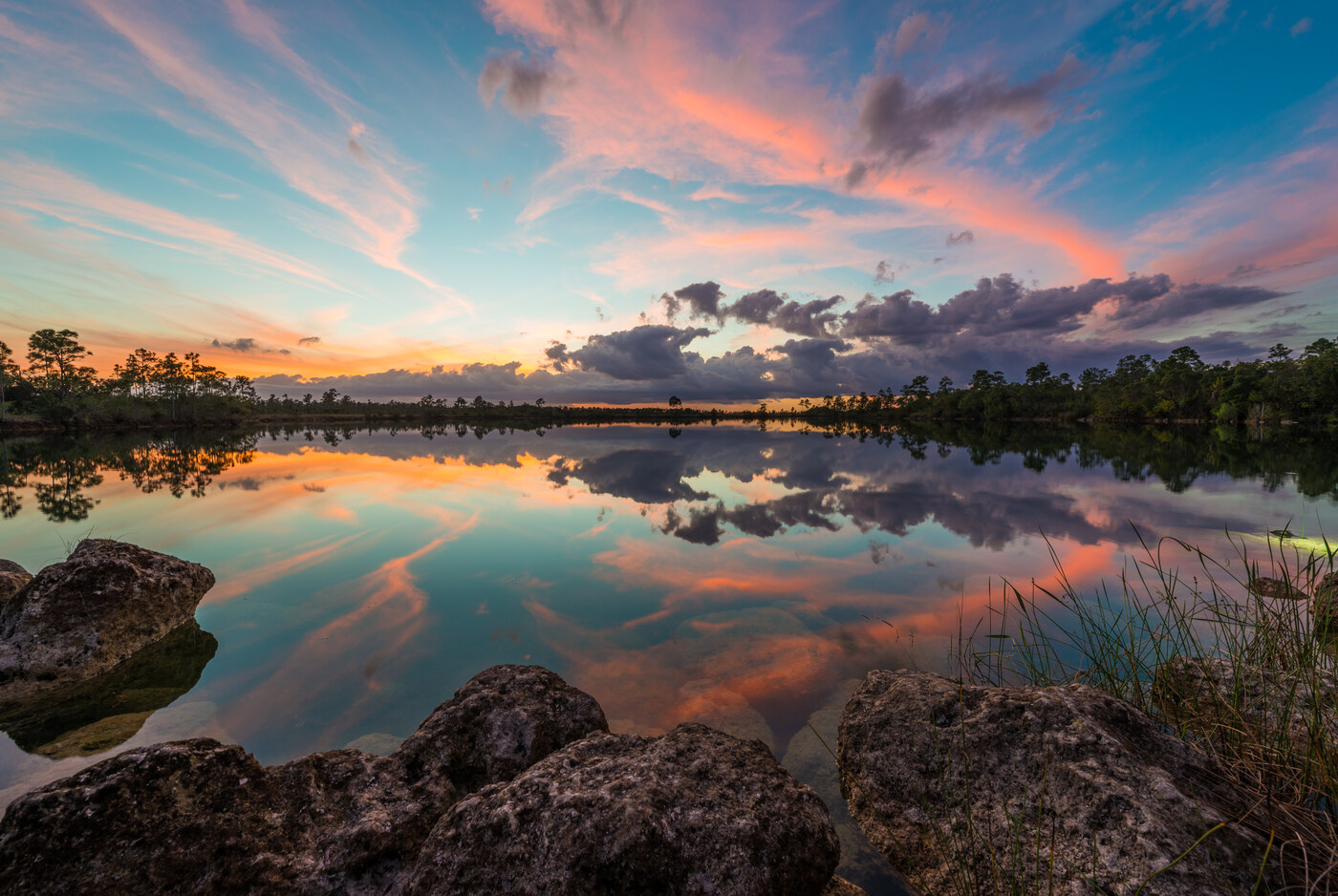A calm lake at dusk reflecting a blue, orange and purple sky, with trees along the far shore, rocks at the left and grass at the right