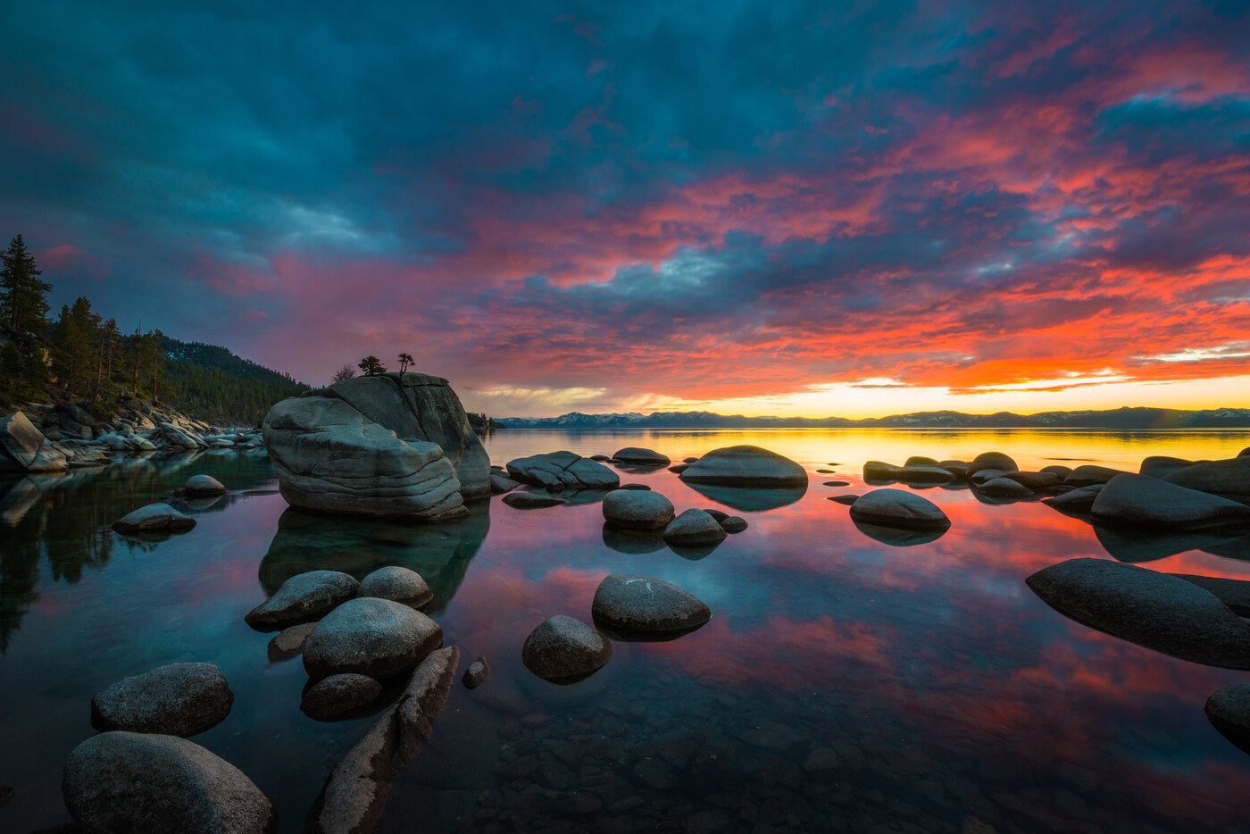 Colorful sunset sky reflected in calm shallow water around dark rounded rocks