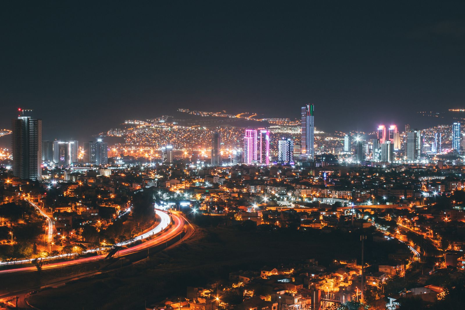 Wide view of a city and hillside covered in glowing lights at night with a light-trail road curving through the scene