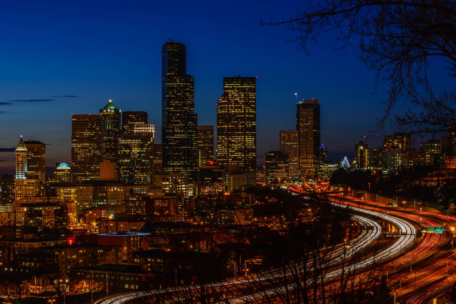 City skyline of lit towers against a deep blue twilight sky with light trails flowing along a highway