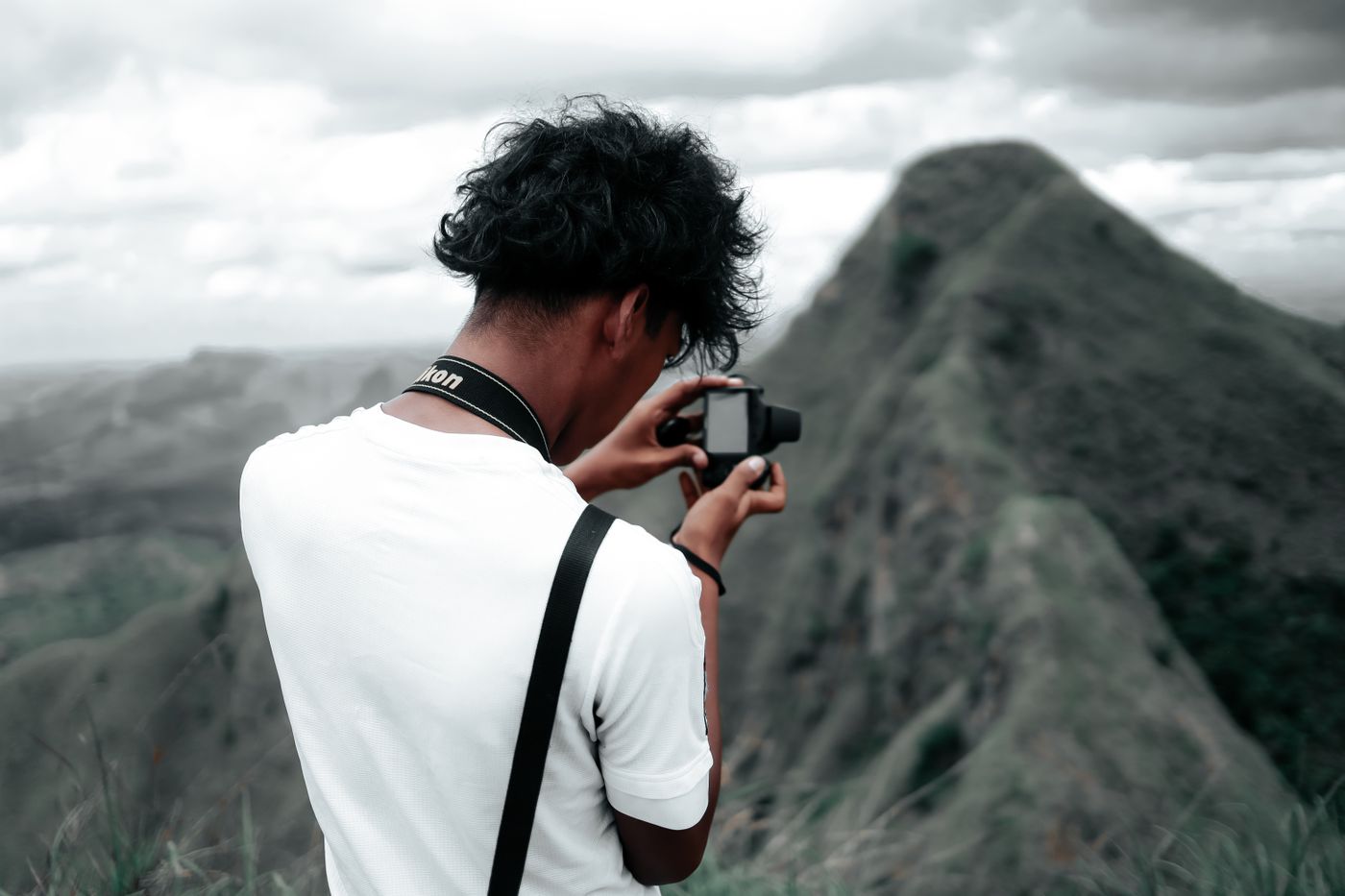 A photographer in a white t-shirt holding a camera in front of a rocky mountain ridge under a cloudy sky.