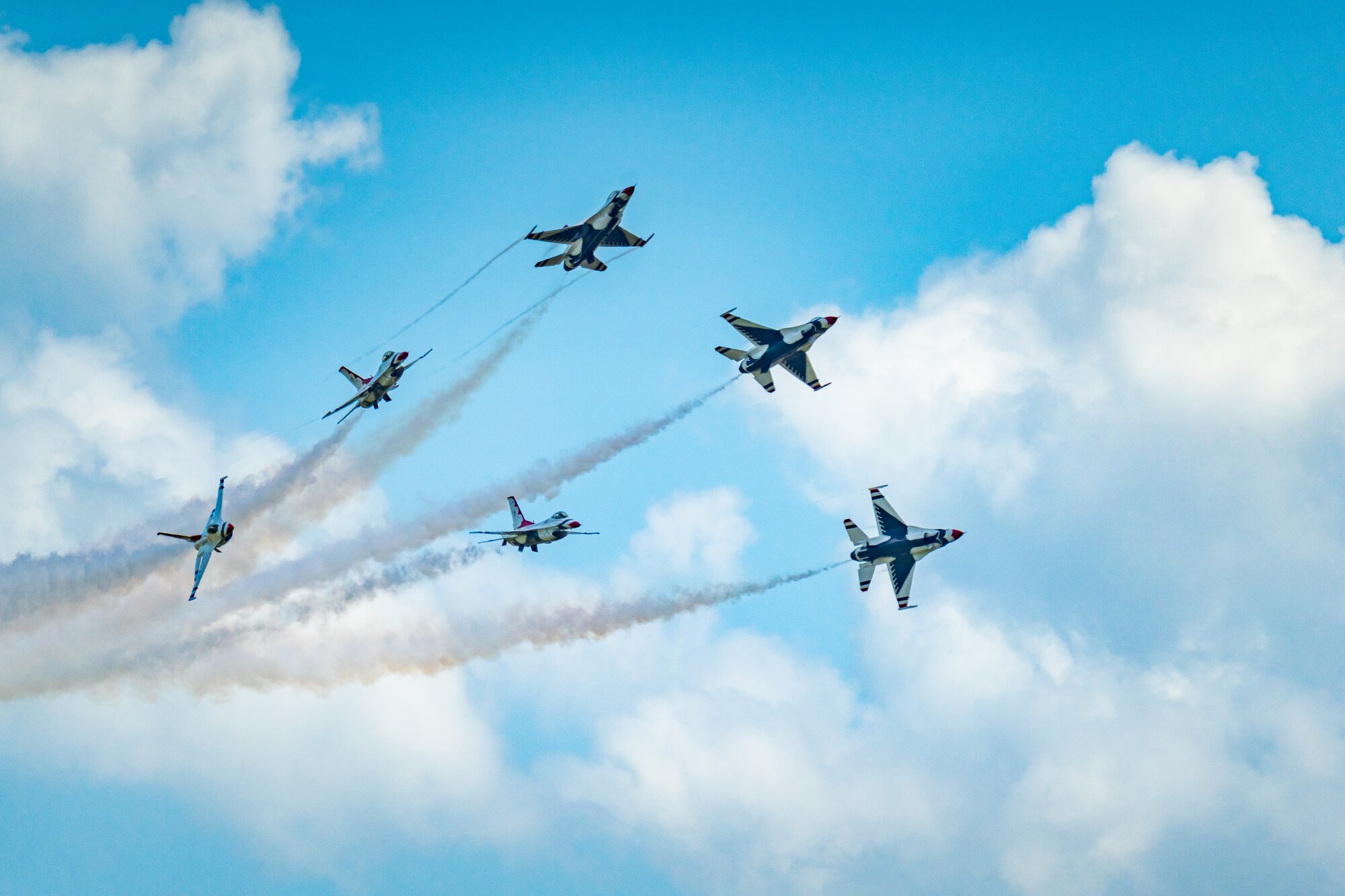 Six jets breaking away from each other with white smoke trails against blue sky and white clouds