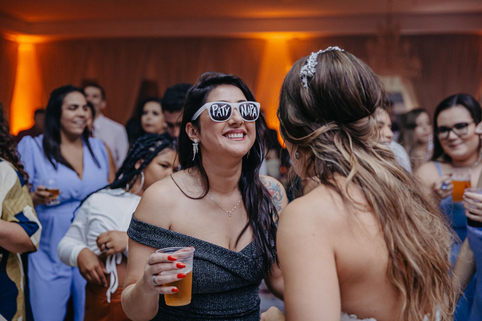 Guests at a warmly lit wedding reception laughing and celebrating, with a woman in sunglasses in the foreground.