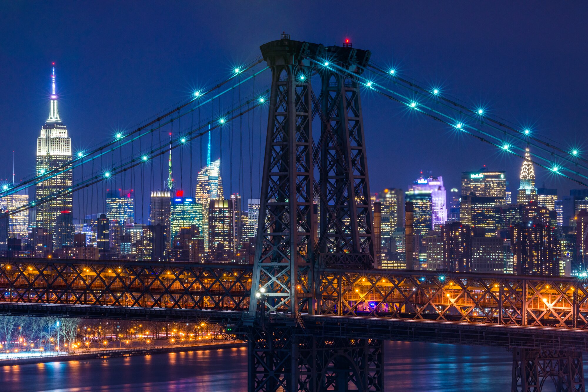 Cityscape photo of a suspension bridge tower with the Midtown Manhattan skyline glowing behind it at night