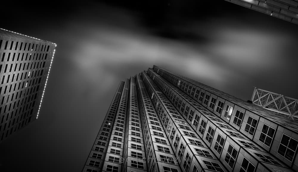 Black and white long exposure photograph looking up at downtown skyscrapers with streaking clouds in a night sky