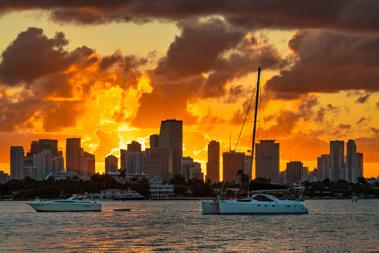 A sailboat and a motorboat on the water in front of the Miami skyline under a fiery orange sky with dark clouds